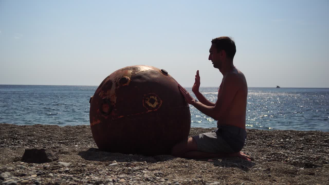 hombre haciendo ejercicio de yoga al aire libre cerca de una vieja mina marina flotante oxidada en la playa con costa rocosa y fondo marino. estilo de vida saludable, contaminación, protección de la naturaleza, concepto de guerra y paz