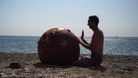 hombre haciendo ejercicio de yoga al aire libre cerca de una vieja mina marina flotante oxidada en la playa con costa rocosa y fondo marino. estilo de vida saludable, contaminación, protección de la naturaleza, concepto de guerra y paz