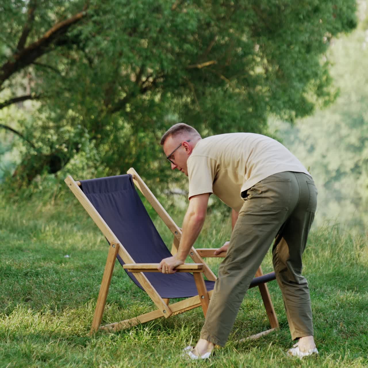 Caucasian man sets folding chair on the grass. Man chooses the best place for the chair. Nature at backdrop