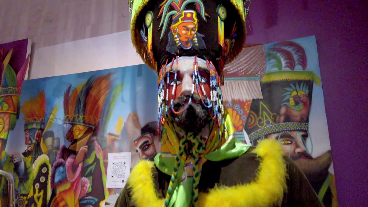 Maschera tradizionale messicana da festival e costume colorato esposti in un museo culturale, con un copricapo elaborato, viso dipinto, perline decorative, piume e opere d'arte sullo sfondo.