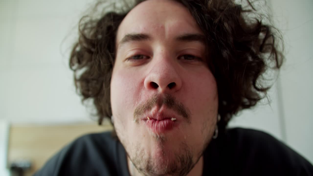 곱슬머리와 수염을 가진 행복한 남자의 클로즈업 초상화가 현대 아파트에서 아침에 햄버거를 고 즐기고 있습니다.