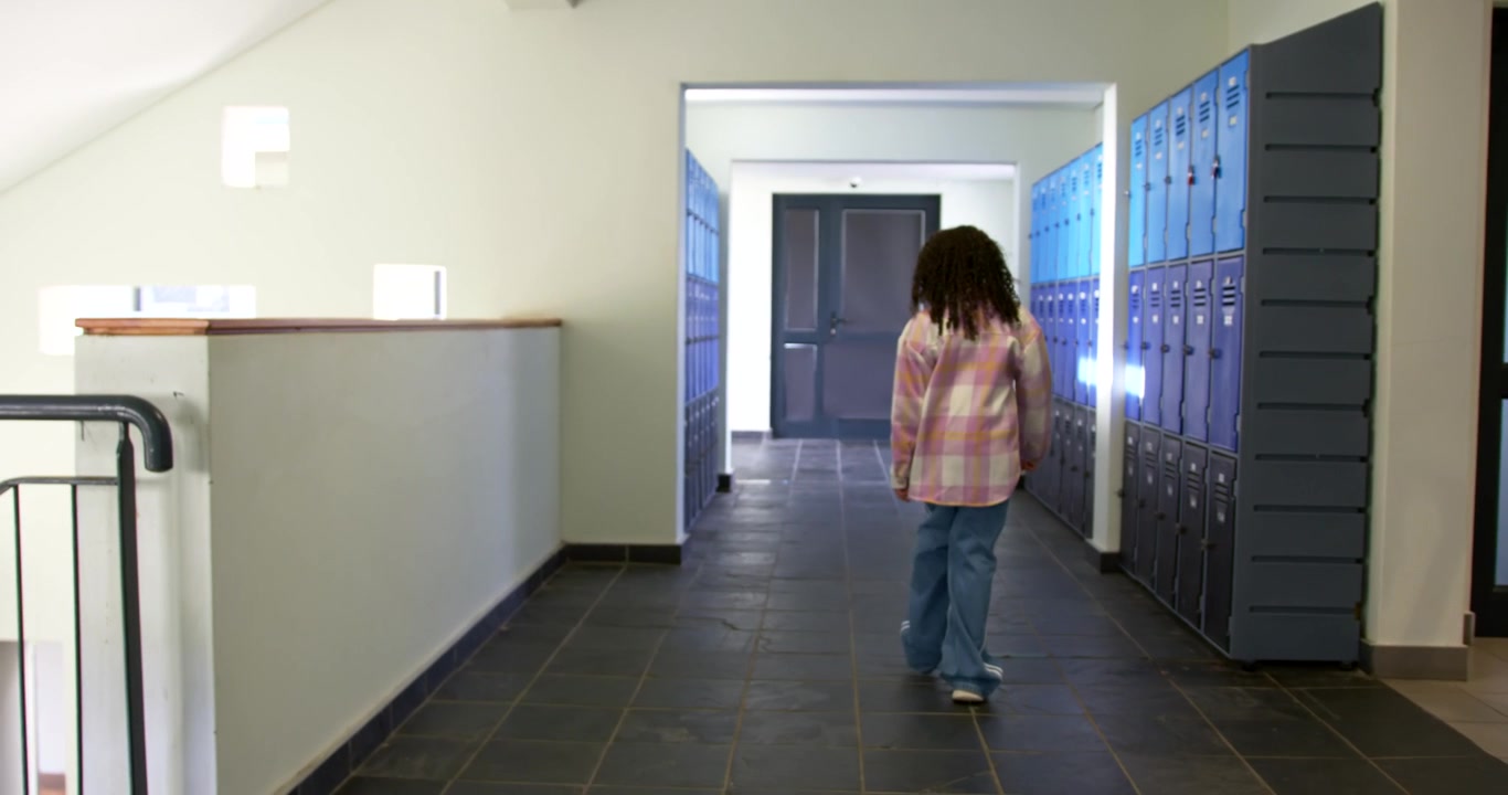 Niña afroamericana en edad escolar caminando por el pasillo de la escuela, pasando taquillas azules, hacia puertas dobles.