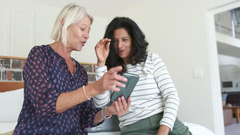 Two-happy-diverse-senior-women-using-smartphone-and-laughing-in-sunny-bedroom,-slow-motion