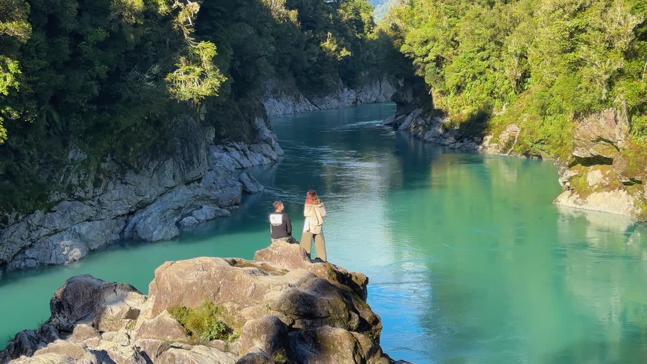 Um casal está sentado em um mirante panorâmico no desfiladeiro de Hokitika, Nova Zelândia, admirando o rio turquesa e a floresta ao redor. A paisagem tranquila e a natureza vibrante criam uma experiência serena.