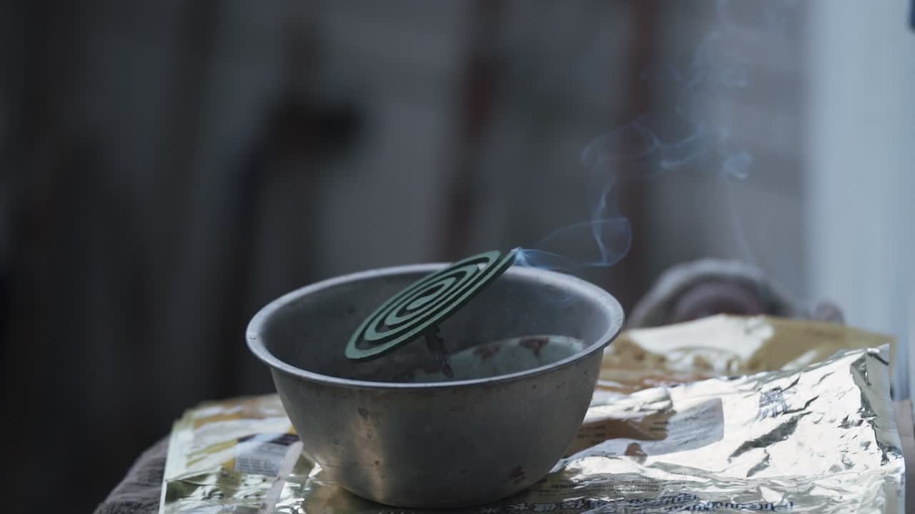 A close-up of a green spiral mosquito coil burning inside a rustic metal bowl, with a steady wisp of smoke rising, commonly used as an insect repellent in tropical climates