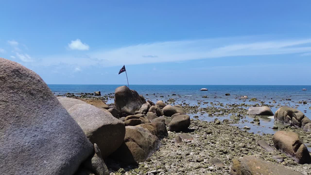 A pirate flag waves on a rocky shoreline at Lamai Beach, Koh Samui, Thailand. Turquoise water, clear blue sky, and scenic coastal landscape