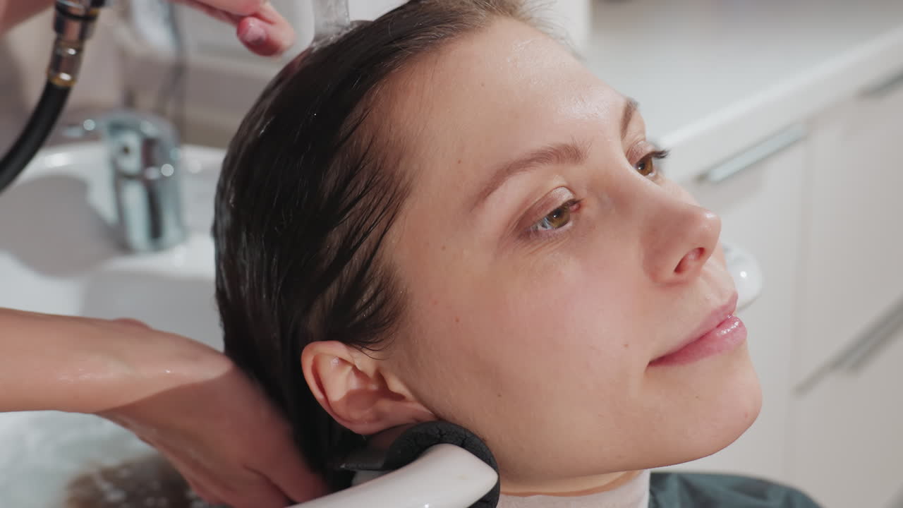 미용 세션 스냅 사진, 여성이 편안한 머리 감김을 받고 따뜻한 물과 부드러운 흐름으로 편안한 살롱 트리트먼트를 받으며, 전문적인 살롱 환경에서 진정 샴푸를 즐기는 여성의 클로즈업 장면