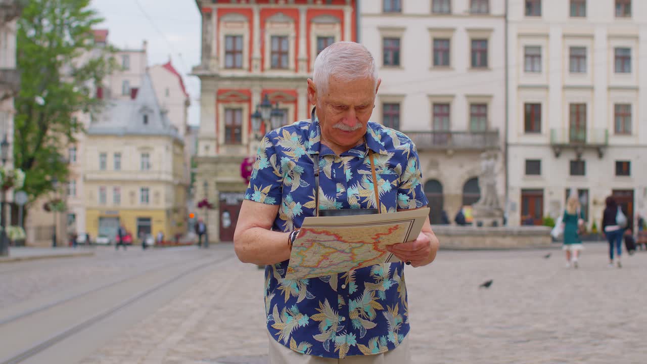 Anziano nonno turista elegante che cammina per strada cercando la via usando una mappa cartacea in città.