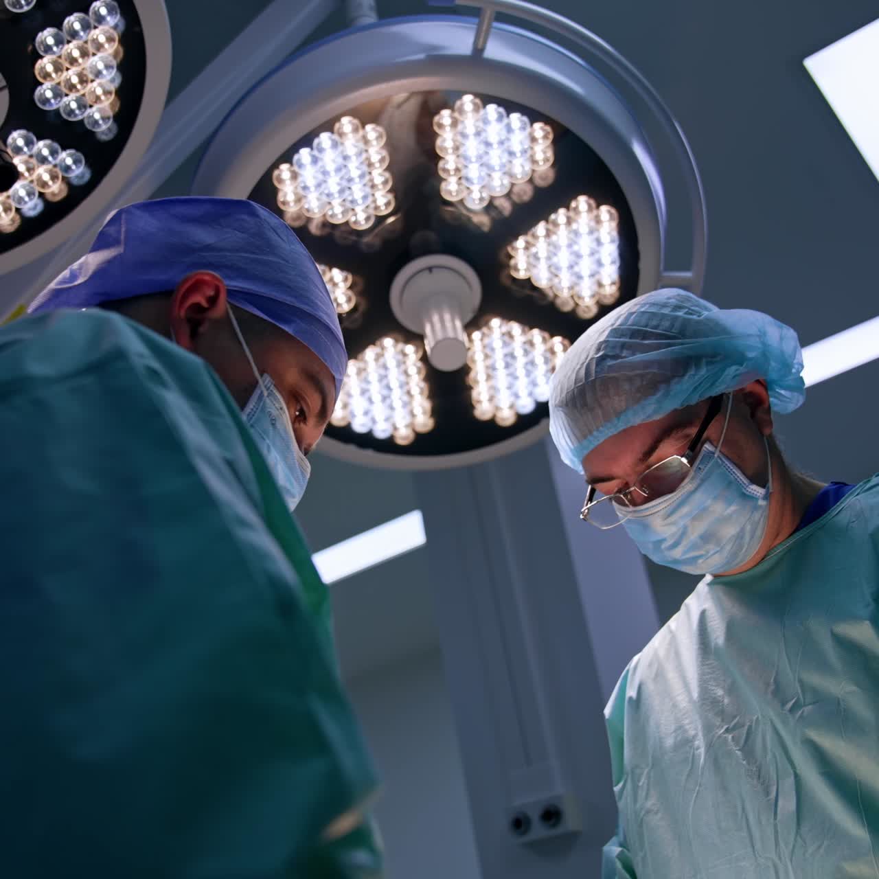 Experienced male surgeons work together at operation. Two big round lamps on the ceiling over the doctors. Low angle view
