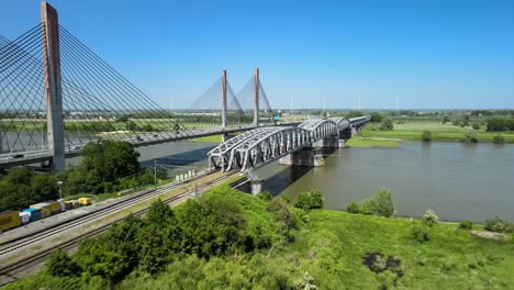 Vista-Aérea-Del-Tren-Holandés-Cruzando-Un-Puente-Ferroviario-En-Un-Día-Soleado-Rodeado-De-Campos-Verdes-Y-Río