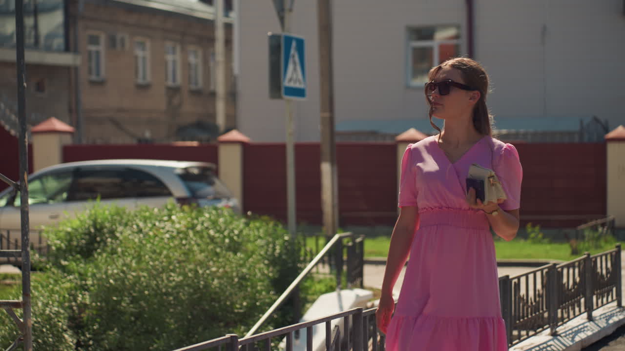 Mulher caminhando na calçada em área urbana, usando vestido de verão rosa e óculos de sol, bebendo café enquanto passa por um parapeito, arbustos e prédios residenciais sob a luz quente do sol, em um ritmo relaxado e com um estilo casual
