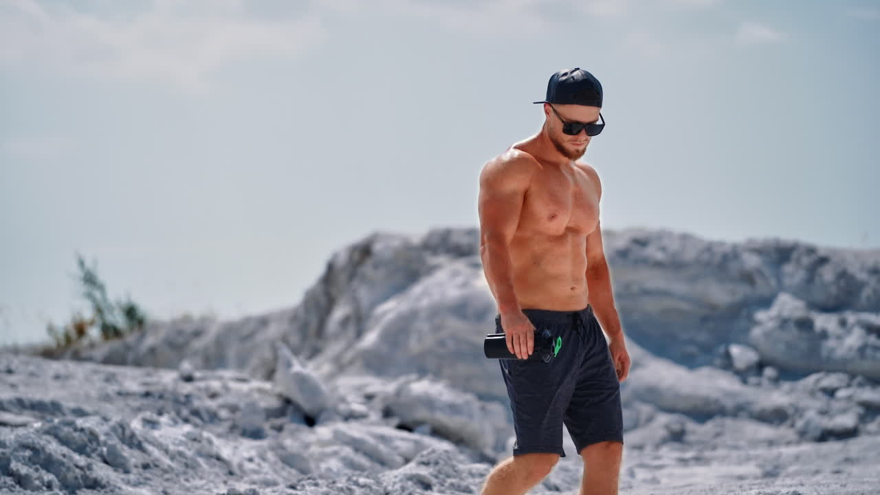 Handsome man with trained body holding flask in the mountains. Muscular athlete in shorts and sunglasses descends from the white hill in a sunny summer day.