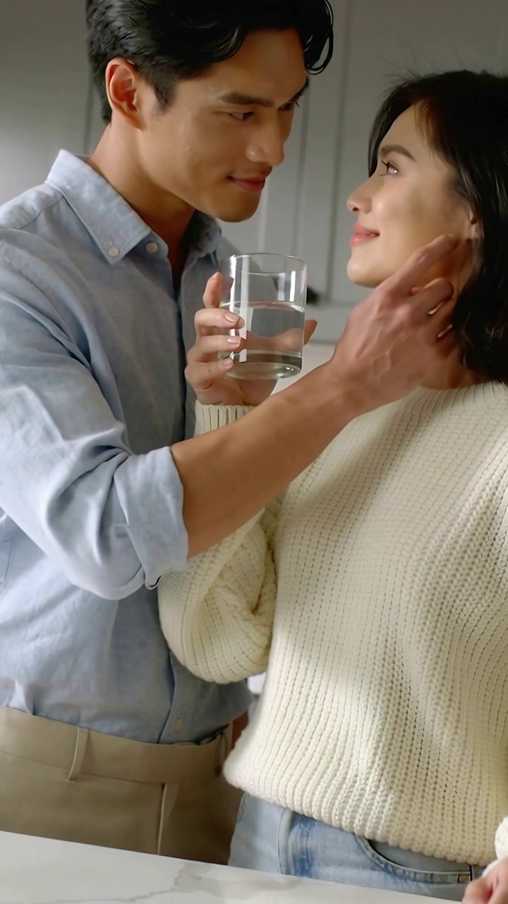 Video verticale: Uomo in camicia blu che si china e abbraccia una donna in cucina dopo un contatto visivo, con un bicchiere in mano.