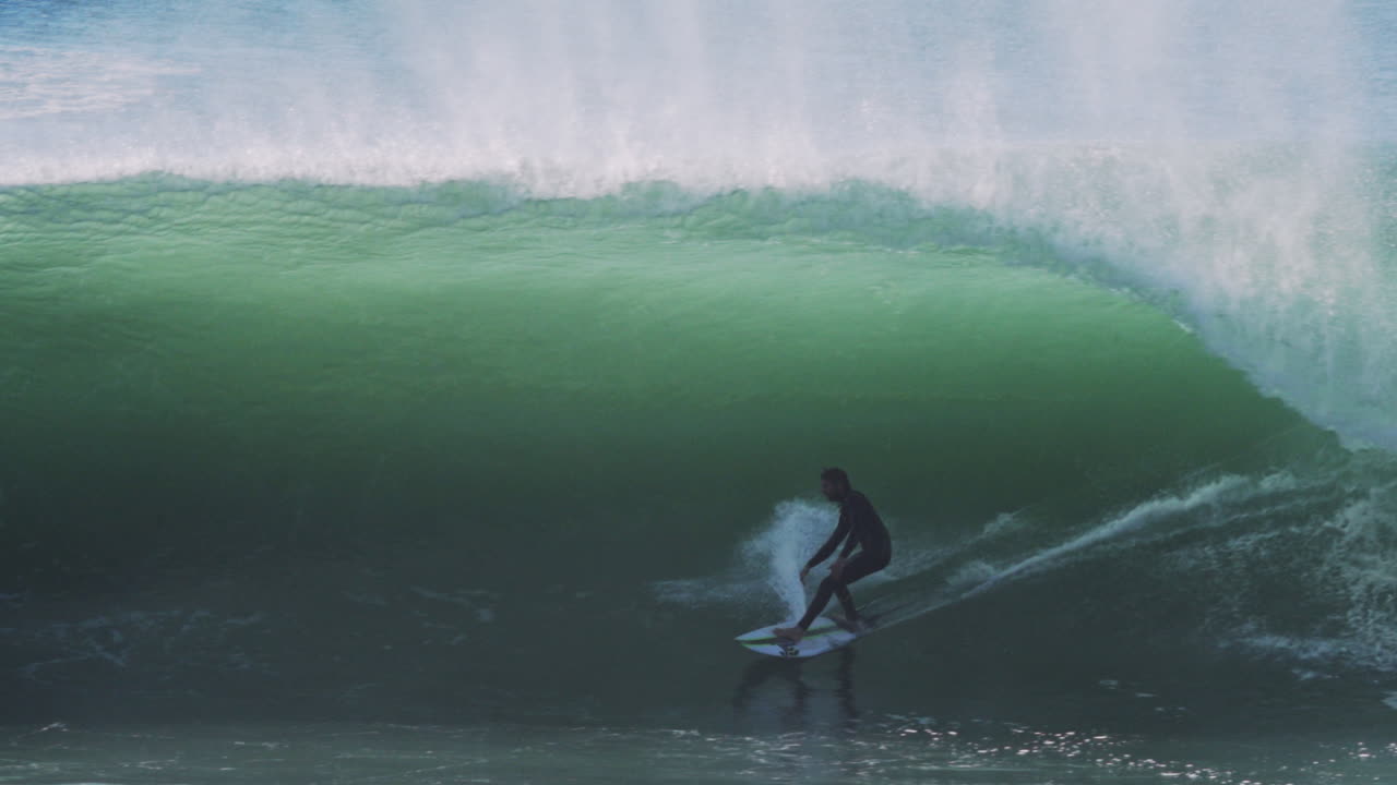 Slow motion surf barrel breaks over rider framed in perfect hollow green tunnel