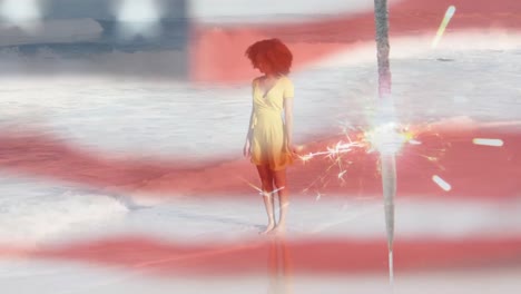 Animation-of-american-flag-waving-and-sparkler-over-woman-standing-on-beach