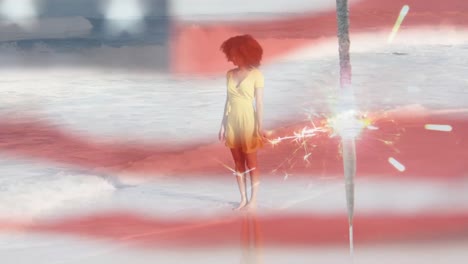 animation of american flag waving and sparkler over woman standing on beach