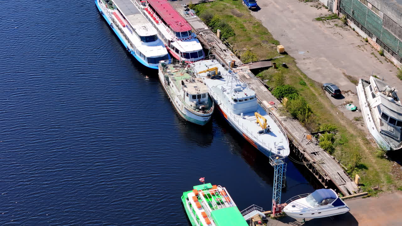 Aerial View of a Busy Riverside or Canal Dockyard Featuring Various Moored Vessels, from Brightly Colored Passenger Ships and Workboats to Smaller Yachts, on a Sunny Day in an Urban Industrial Setting