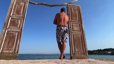 puerta de madera decorativa frente al mar y vista trasera del hombre de pie en traje de baño apoyado contra el marco de la puerta