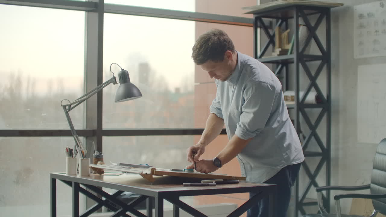 мужчина - молодой современный архитектор, сидящий за столом, рисует план здания и разрабатывает дизайн, сидя в офисе на чердаке рядом с большим окном, чтобы создать фасады зданий, проектировать мебель и проектировать проект жилого комплекса.