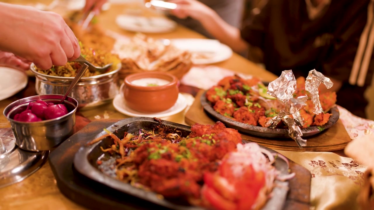 Close-up of hands using tongs to serve spicy Indian food on a crowded dinner table.