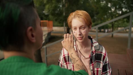 over the shoulder: a girl with a short haircut with green hair in a green shirt applies glitter with her finger to the face of her girlfriend with short orange hair in a checkered pink shirt on brown benches in the park