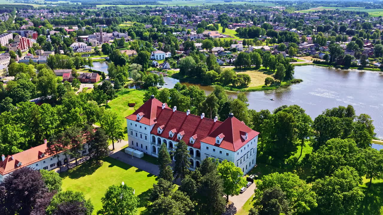 Aerial reveal shot of historic Biržai Castle with red roof, nestled in lush parkland beside a tranquil lake in Biržai, Lithuania