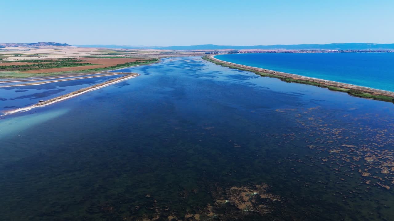 En drone flyr over grunt kystvann og en smal landtunge som deler sjøen nær landlige Pomorie-landskap.