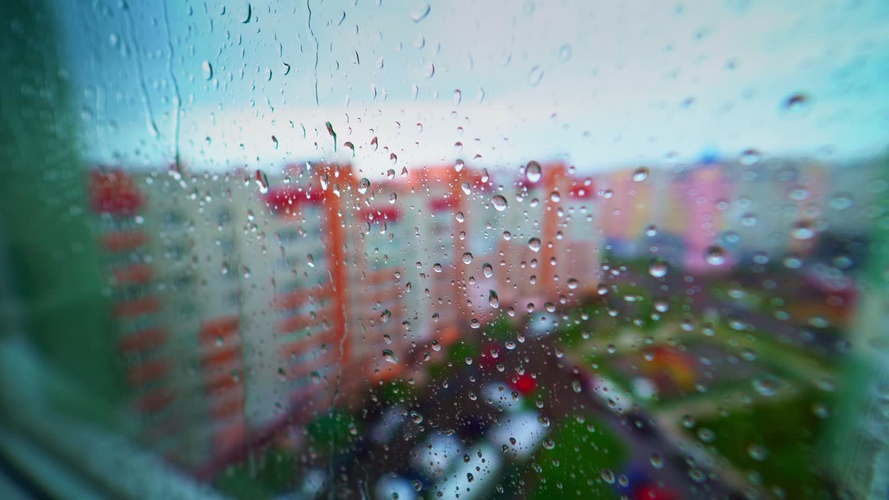 Rain droplets on window pane. View from the window on rain falling outside on colorful city buildings background. Rainy weather.