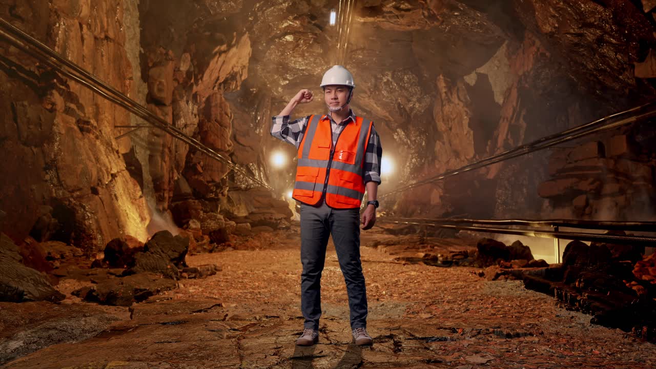 Full Body Of Asian Male Engineer With Safety Helmet Flexing His Bicep And Smiling To Camera While Standing In Underground Mine Tunnel