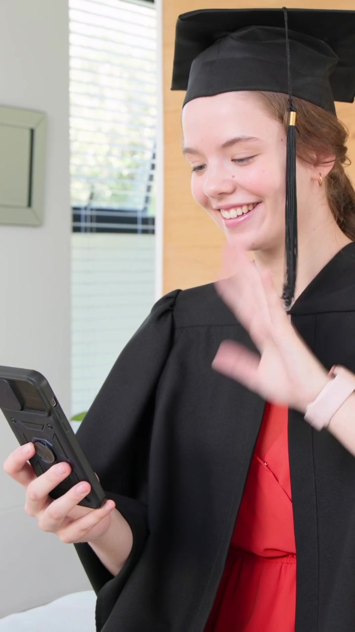Video verticale: Neolaureato sorridente saluta con la mano e poi controlla lo smartphone in una stanza, indossando tocco, toga e nappa.