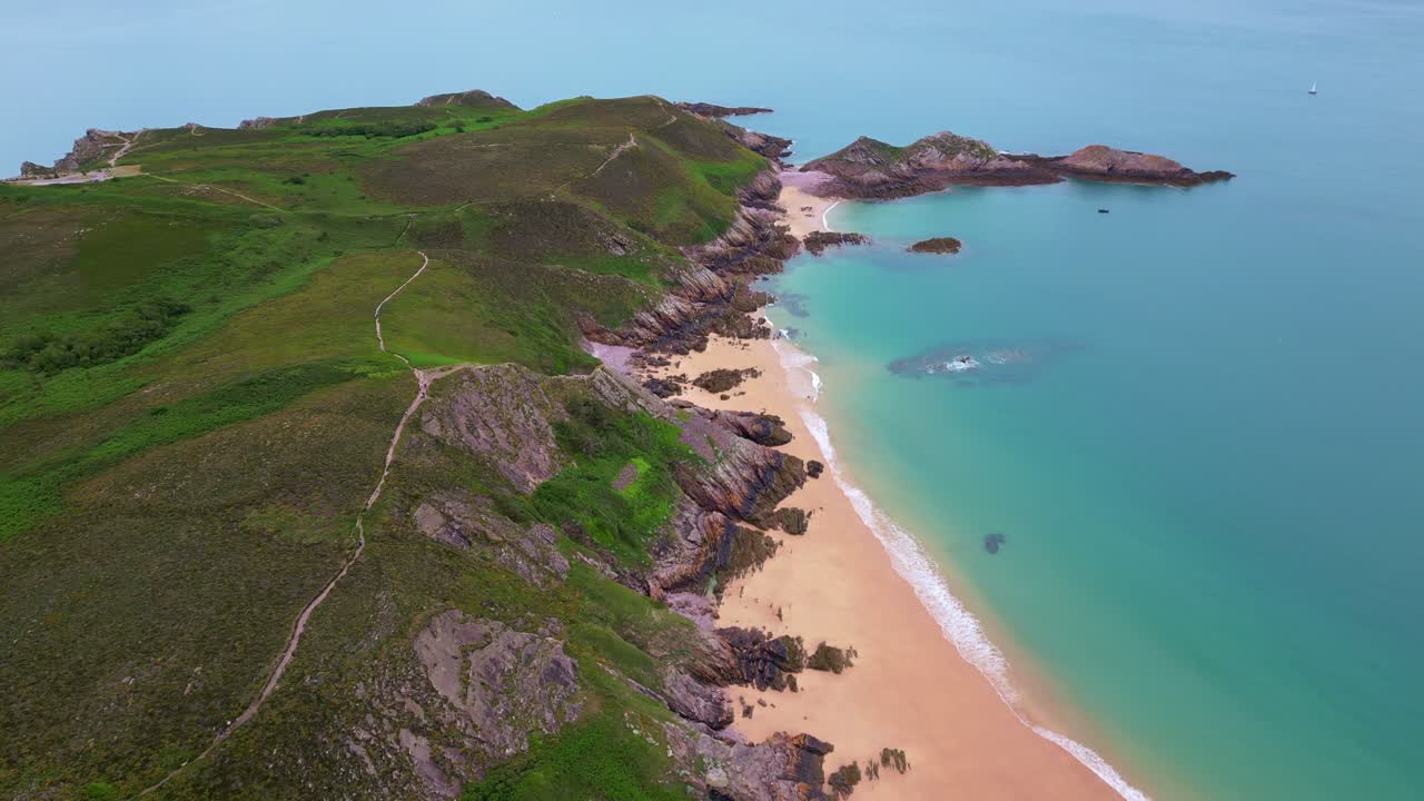 Aerial drone view of the scenic Cap d'Erquy peninsula with lush green heathland, rocky coast, sandy beach, and turquoise water, Brittany, France