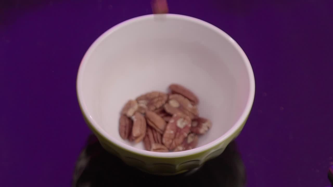 Pecan nuts fall into a yellow bowl in fast motion against a purple background.