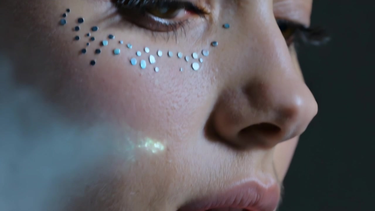 Blinking model posing on camera cue, lowering gaze for studio beauty shoot, with shiny face gems