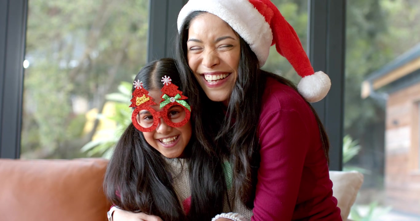 Madre e hija diversas abrazándose y riendo después de que la madre le pusiera gafas de Navidad a la niña en el sofá.