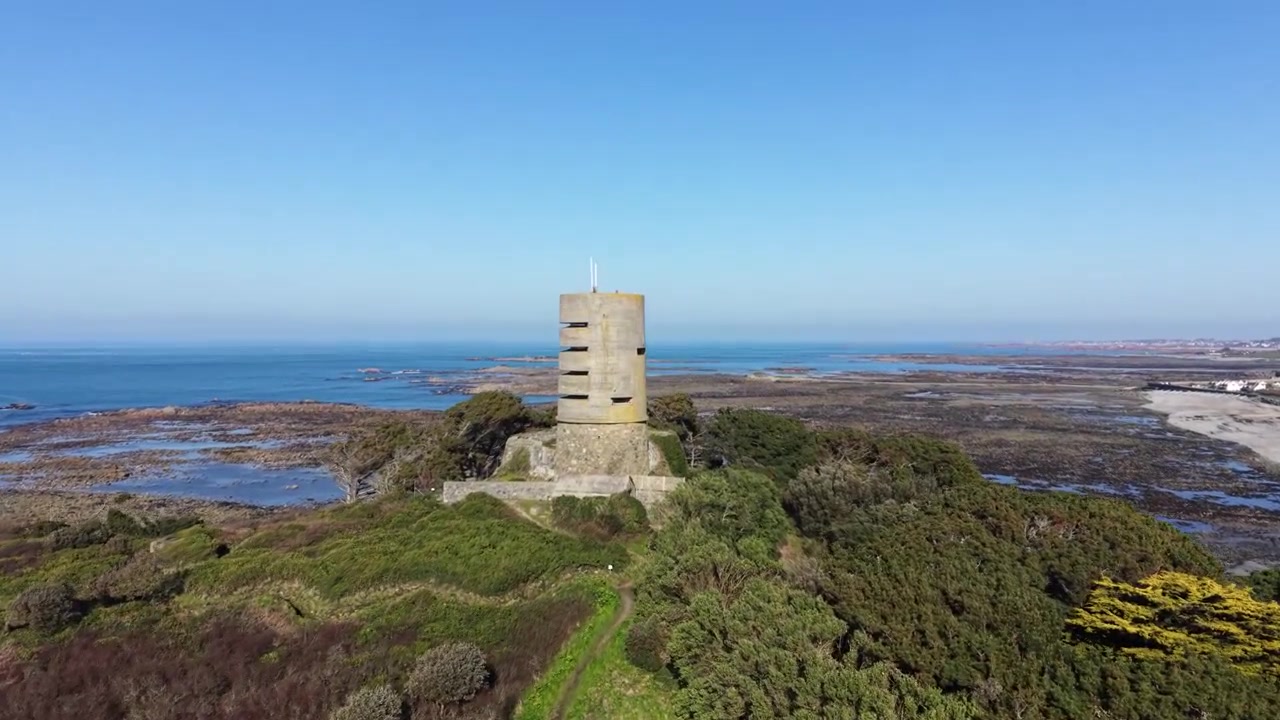 L’Eree, Guernsey: Öğleden sonra güneşinde, çarpıcı berrak mavi gökyüzüne karşı denize ve Lihou Adası'na bakan burun üzerindeki eski bir Alman gözetleme kulesinin dramatik, ileriye doğru yükselen drone görüntüleri