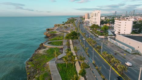 Drohnenflug-Entlang-Der-Küstenstraße-Des-Bezirks-Malecon-Und-Verkehr-Zur-Goldenen-Stunde---Wunderschöne-Landschaft-In-Der-Stadt-Santo-Domino,-Dominikanische-Republik