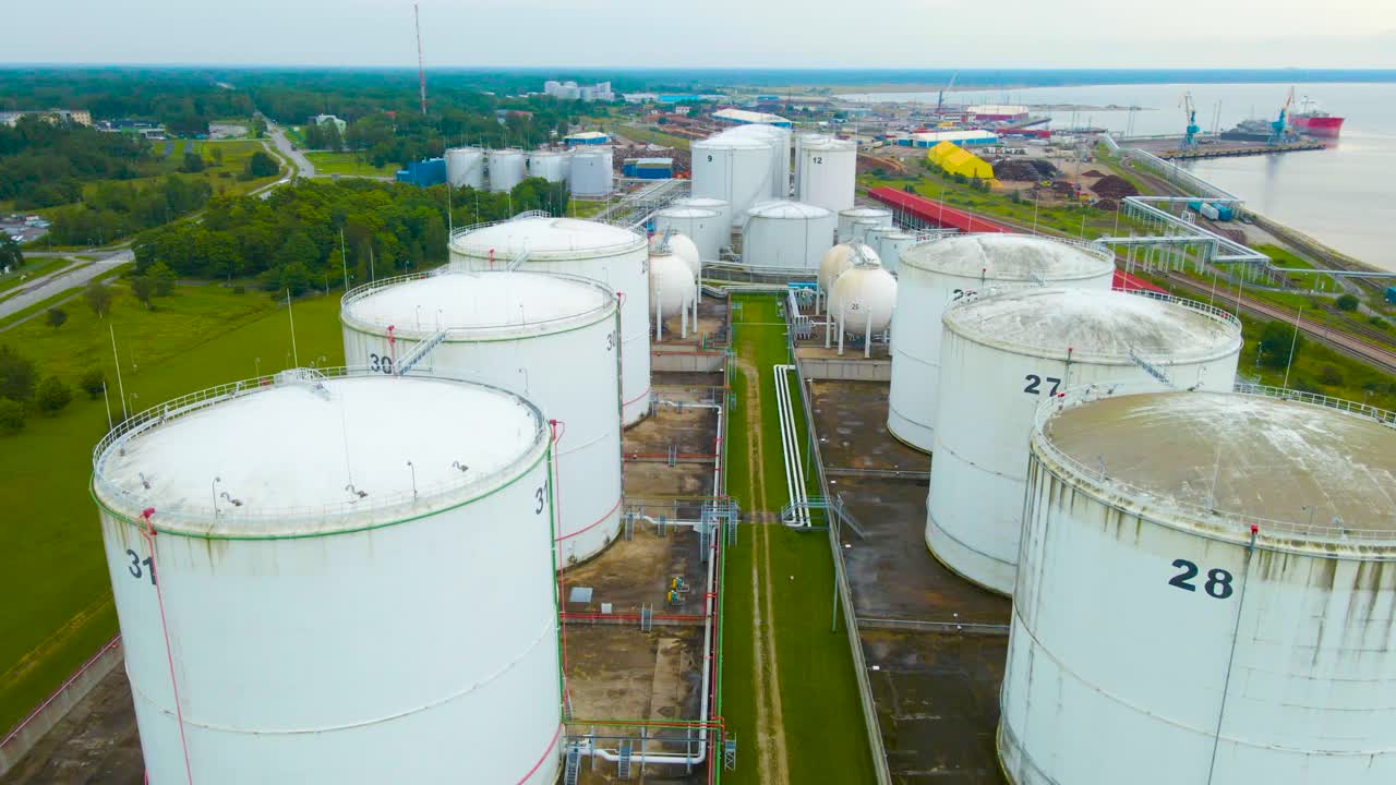 Aerial frone footage flying over and gliding above large white colored liquid strorage tanks near a harbor and sea shoreline where a railroad is visible also during summer cloudy day. Numbered tanks