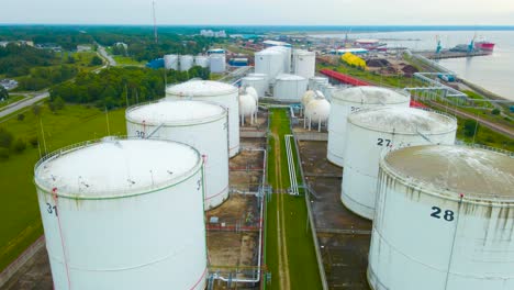aerial frone footage flying over and gliding above large white colored liquid strorage tanks near a harbor and sea shoreline where a railroad is visible also during summer cloudy day. numbered tanks