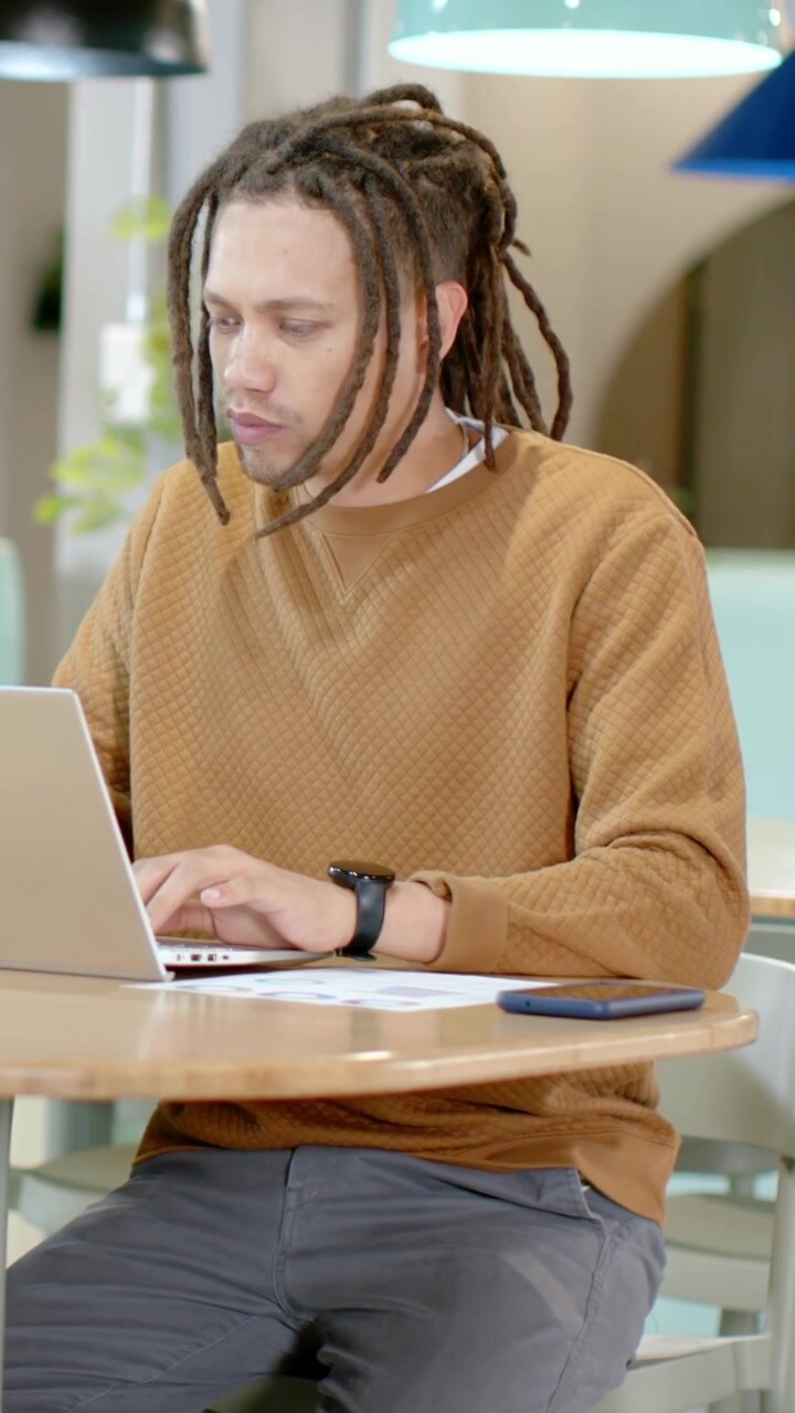 Vertikal video: Mand i brun sweater starter arbejde, taster på bærbar computer på en café med smartphone og papir.
