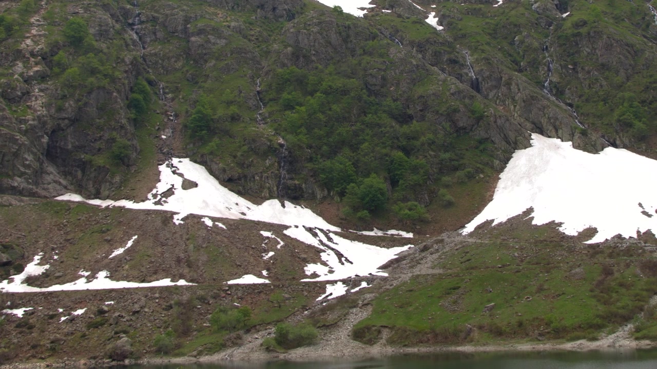 Vidvinkelbilde av steinete fjellsider med grønn vegetasjon, snøflekker og en foss
