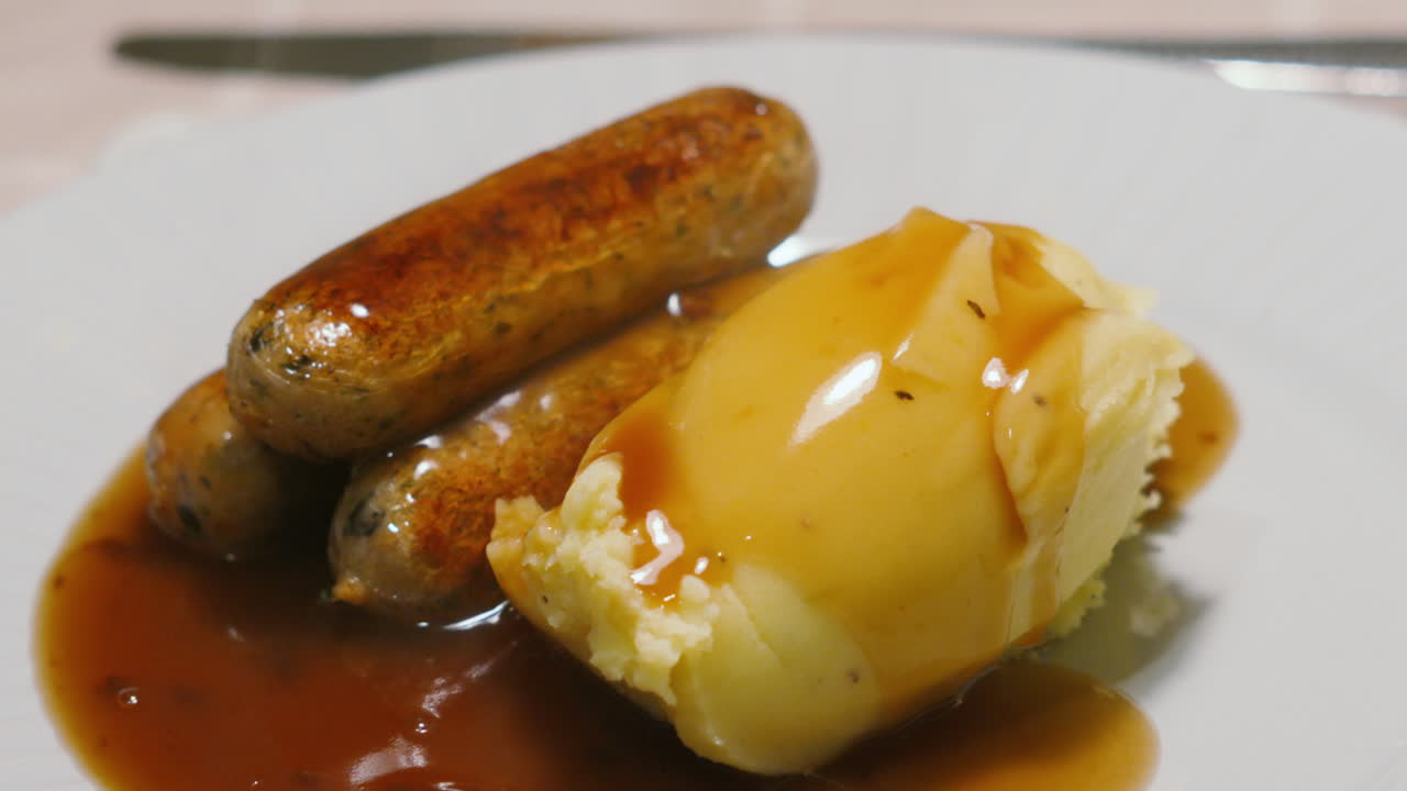 Primo piano al rallentatore del vapore che si sprigiona da salsicce fresche e purè di patate cremoso su un piatto bianco. Cinematografia alimentare di alta qualità per menù invernali