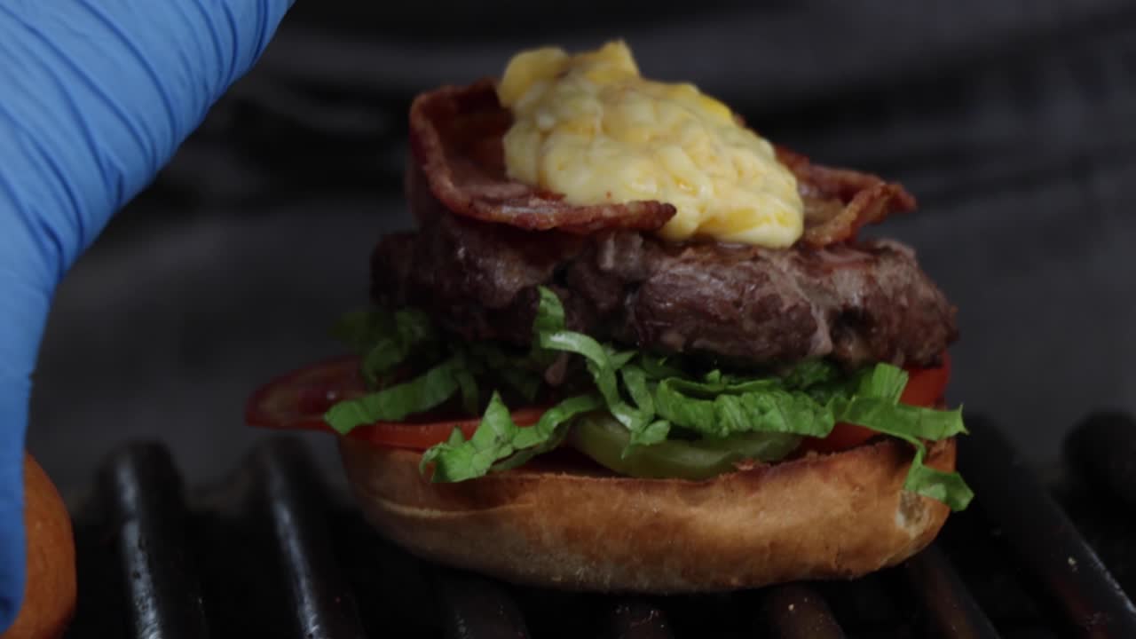 un chef terminando de montar una hamburguesa a la parrilla con carne, tocino, queso, tomate, lechuga y pan entero