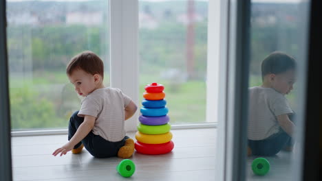 baby boy assembled the colorful toy pyramid. kid wants to take the top but distracts on something and moves back.