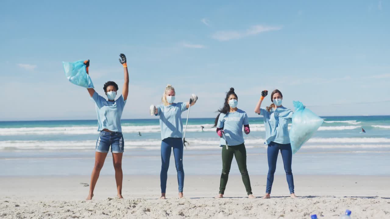 자원 봉사자 티셔츠와 얼굴 마스크를 입은 다양한 여성 그룹이 해변에서 쓰레기를 집어 들고 있습니다.