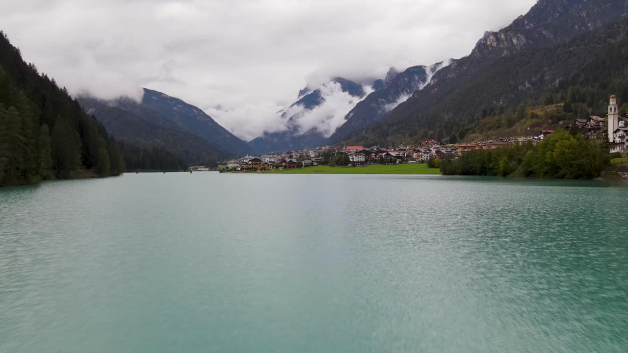 volando sobre el lago azul hacia un pequeño pueblo rodeado de montañas en un día nublado