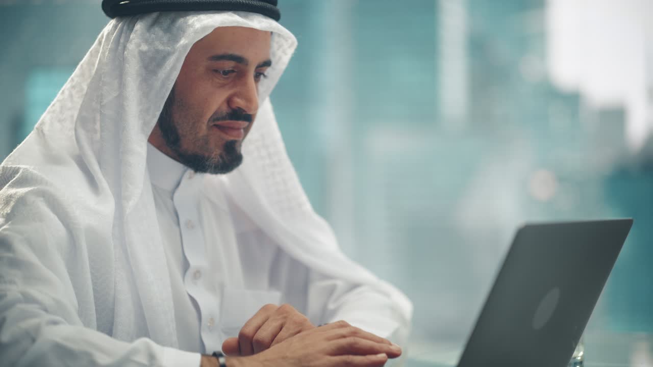 empresario árabe en traje tradicional blanco sentado en la oficina y trabajando en una computadora portátil. gerente de negocios celebra un acuerdo de inversión exitoso. saudi, emirati, concepto de empresario árabe.
