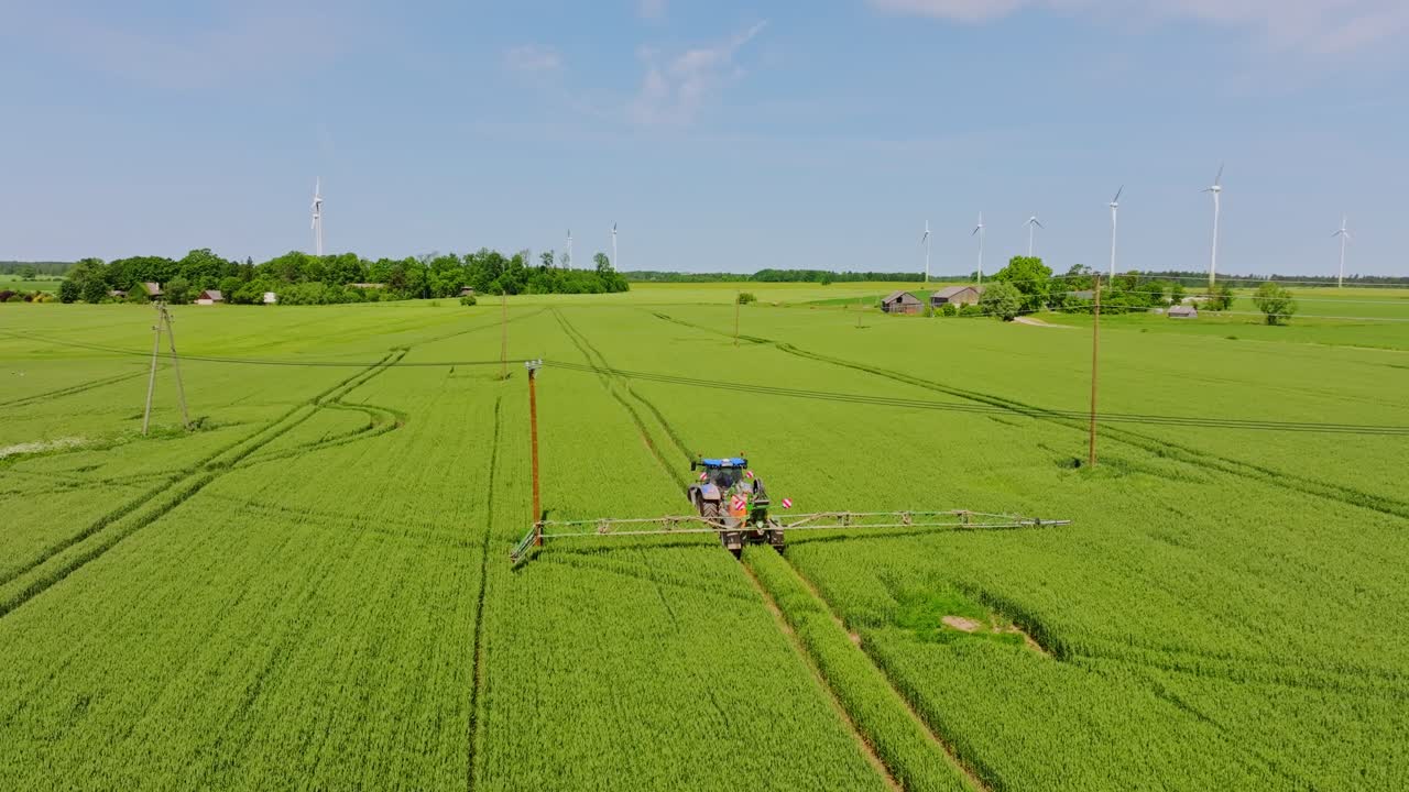Cinematic rural view of tractor spraying farmland, Latvia, wind energy landscape