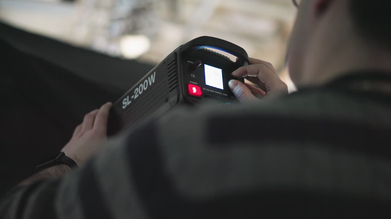 camera technician adjusting brightness knob and power switch on SL-200W LED studio light mounted on stand, manipulating control panel with cables and blue backdrop during broadcast production