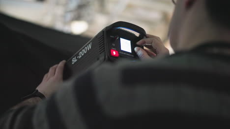 camera technician adjusting brightness knob and power switch on sl-200w led studio light mounted on stand, manipulating control panel with cables and blue backdrop during broadcast production