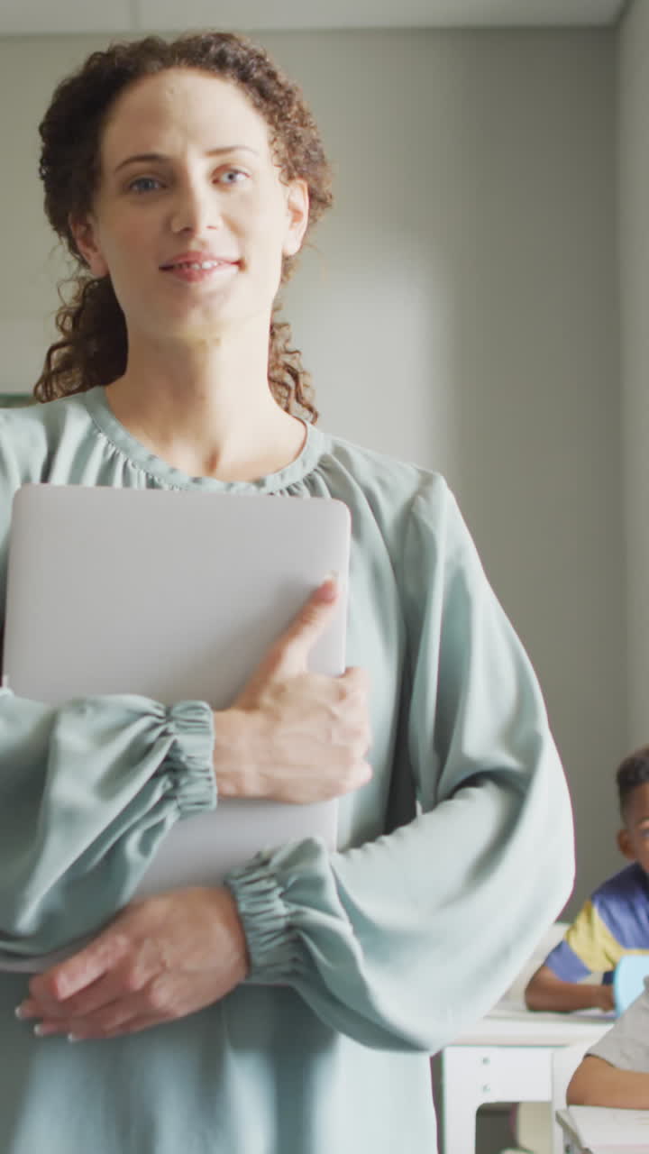 video de una feliz maestra caucásica sosteniendo una computadora portátil en una clase de alumnos diversos